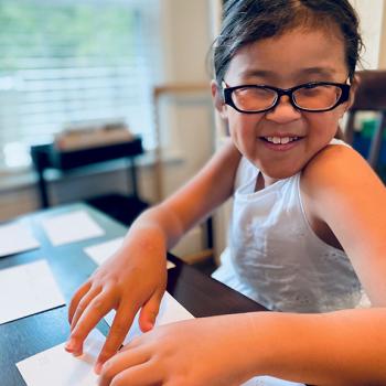 A young girl smiles widely as she reads Braille off of a notecard